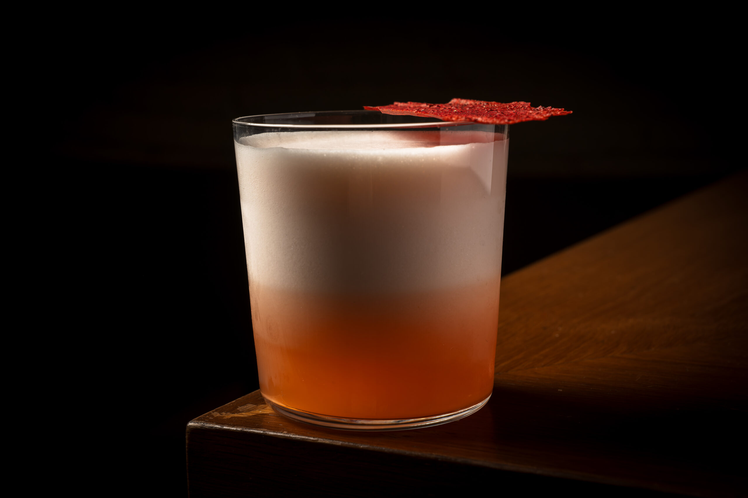 Royal sour de pisco acholado y aperol en vaso bajo, con espuma blanca, sobre la barra de madera